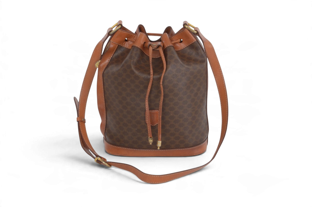 A brown monogram bucket bag with tan leather trim and a matching adjustable shoulder strap, shown on a clean white background.