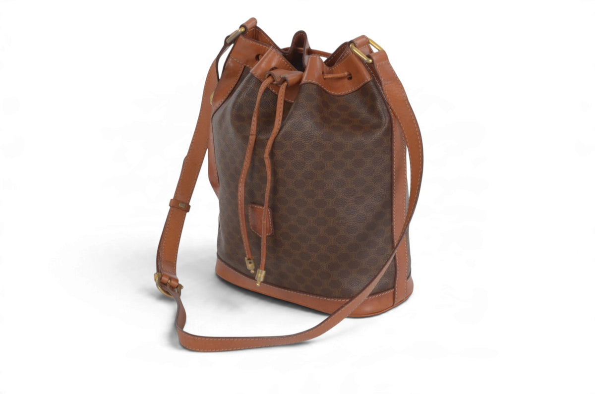 A brown monogram bucket bag with tan leather trim and a matching shoulder strap, shown on a plain white background.