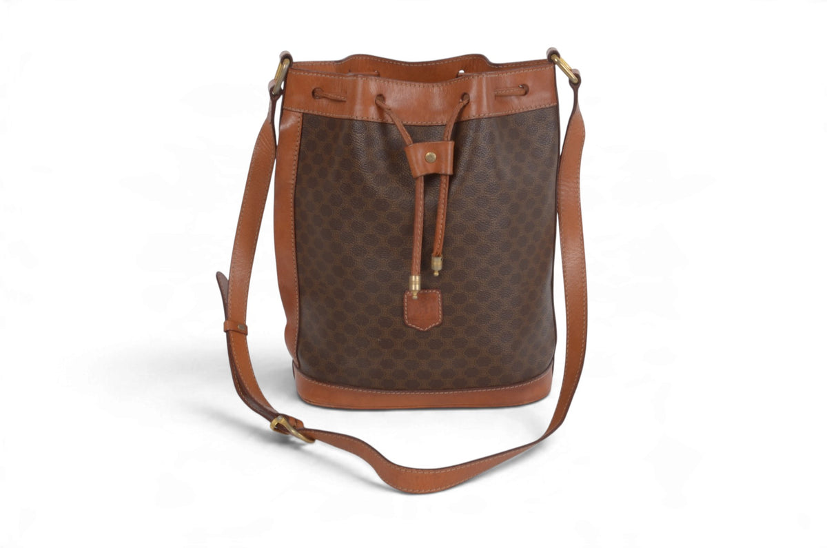A brown monogram bucket bag with tan leather trim and a long adjustable shoulder strap, photographed on a clean white background.
