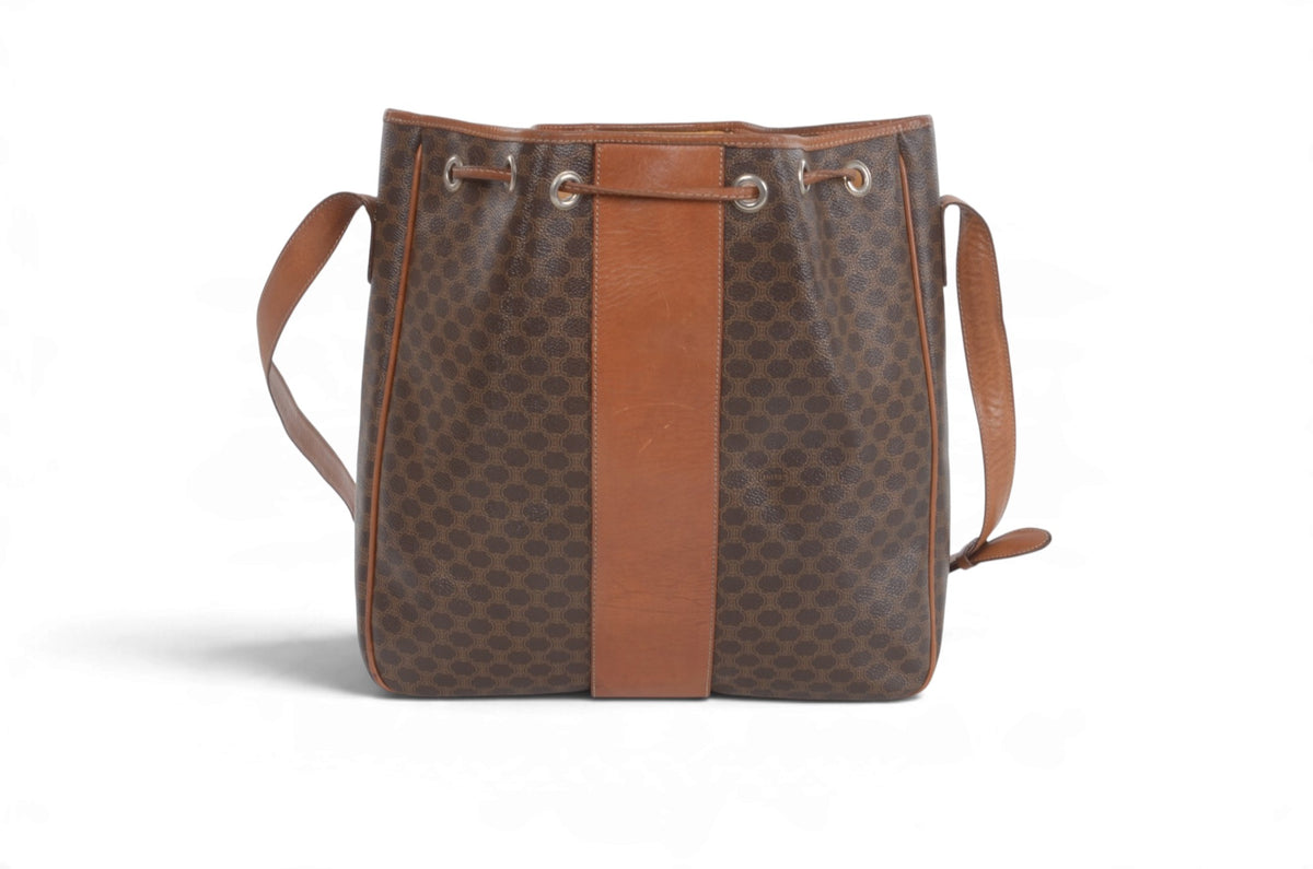 A brown patterned canvas bucket bag with a vertical tan leather stripe down the center, brown leather shoulder straps, and silver-tone metal eyelets at the top, shown on a white background.