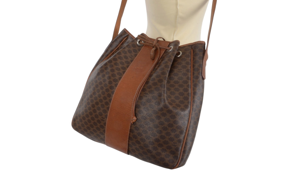 A brown patterned shoulder bag with a central brown leather strap and drawstring closure, displayed on a cream dress form against a white background.