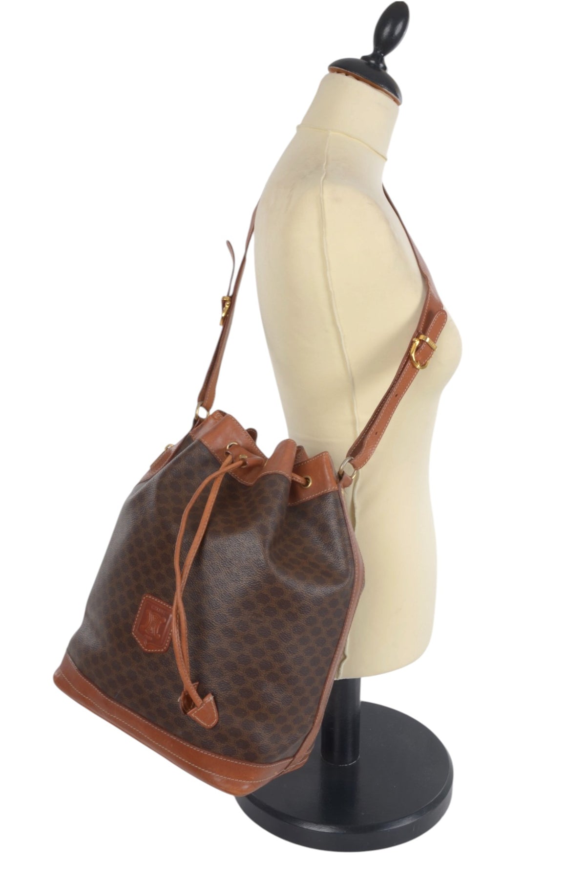 A brown monogram bucket bag with tan leather trim and a drawstring closure, displayed on a cream-colored dress form mannequin against a white background.